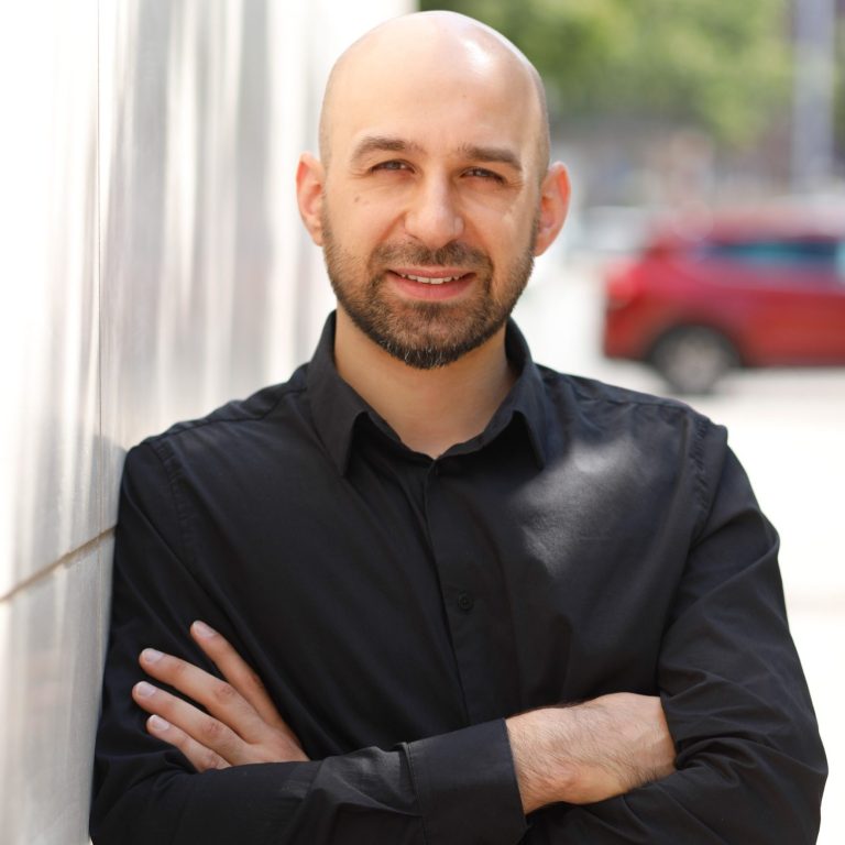 Mann mit kurzem Bart, schwarzem Hemd und verschränkten Armen steht vor einer Wand.